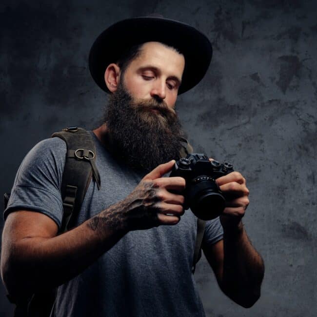 Handsome bearded traveler in a hat with a backpack and tattooed arms, holds a photo camera